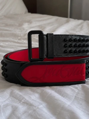 Louboutin belt herr - Snyggt svart bälte i skinn med svarta nitar och en röd insida samt röd detalj med präglad text. Bältet har en stilren, matt svart spänne och cool design som sticker ut.