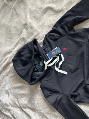 Svart hoodie från Polo Ralph Lauren - Snygg svart hoodie från Polo Ralph Lauren med klassisk röd logga broderad på bröstet. Hoodien har vita dragsnören och en stor huva. Tillverkad i mjukt material som känns skönt mot huden. Perfekt för en avslappnad och stilren look.
