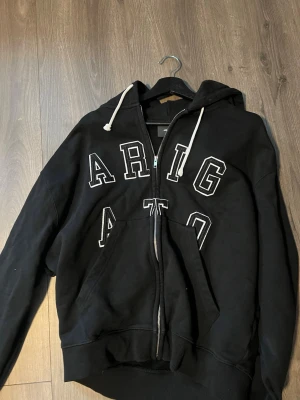 Svart hoodie med dragkedja och tryck - Svart hoodie med vit text 'ARIGATO' i stora bokstäver på bröstet. Modellen har huva med vita snören, dragkedja framtill och en stor magficka. Perfekt för en avslappnad streetwear-look.