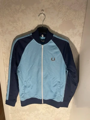 Fred Perry zip tröja  - Sportig ziptröja från Fred Perry i ljusblått och marinblått med vita detaljer. Tröjan har dragkedja, två fickor och ribbade muddar. Fred Perry-logga broderad på bröstet och klassisk retrostil.