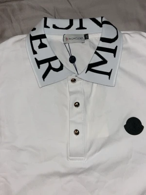 Vit Moncler polotopp med logga - Säljer en vit polotopp från Moncler med stor svart logga på bröstet och en krage med Moncler-text i svart. Toppen har tre knappar framtill och är i ett mjukt bomullsmaterial. Perfekt för dig som gillar stilrena och exklusiva plagg.