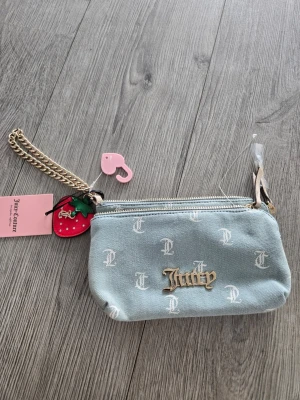 Ljusblå plånbok Juicy Couture - Trendig ljusblå plånbok från Juicy Couture i denimliknande material med vita JC-loggor och guldfärgad Juicy-text framtill. Plånboken har dragkedja, guldfärgad kedja och en söt jordgubbsberlock. Perfekt accessoar för dig som gillar detaljer!