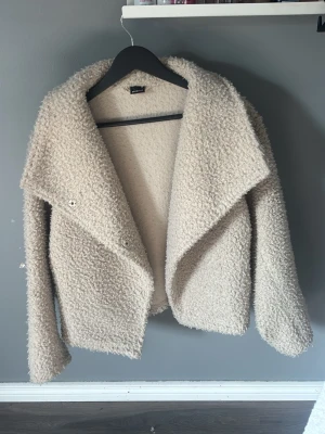 Beige teddyjacka med bred krage - Säljer en supermjuk och fluffig teddyjacka i beige med bred, öppen krage och långa ärmar. Jackan har en oversized look och stängs med knappar framtill. Perfekt för dig som vill ha en cozy och trendig stil i höst och vinter.