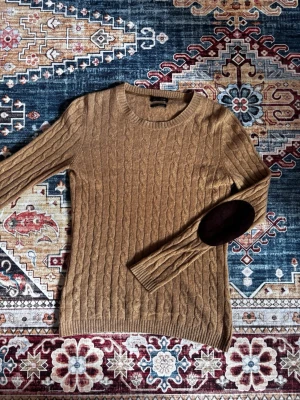 100% Kashmir Tröja från Massimo Dutti - 100% Kashmir Kabelstickad beige tröja med rund hals och bruna mockalappar på armbågarna. Tröjan har långa ärmar och ribbade muddar vid ärmslut och nederkant. Perfekt för dig som gillar klassisk och stilren look.