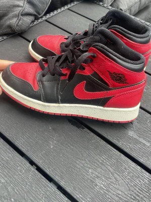 Svarta och röda Nike Air Jordan 1 Mid - Säljer ett par Nike Air Jordan 1 Mid sneakers i svart och rött läder med klassisk röd swoosh och svart snörning. Skorna har vit mellansula och röd yttersula. Ikonisk Air Jordan-logga på sidan. Perfekta för dig som gillar streetwear och snygg design.