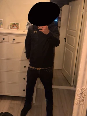 Svart vindjacka från Under Armour - Snygg svart vindjacka från Under Armour med dragkedja framtill och huva med dragsko. Jackan har en minimalistisk design med logga på bröstet och är tillverkad i lätt, vindtätt material. Perfekt för en sportig och stilren look.