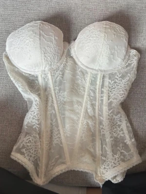 Vit spetskorsett med bygel - Säljer en elegant vit korsett med bygel och vadderade kupor. Korsetten är täckt av vacker spets med blommönster och har hakknäppning bak. Materialet är genomskinligt och ger en romantisk känsla. Perfekt för dig som vill ha något extra snyggt under kläderna.