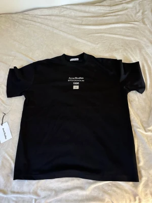 Svart Acne Studios t-shirt - Snygg svart t-shirt från Acne Studios med tryck på bröstet där det står 'Acne Studios STOCKHOLM 1996' och deras ikoniska ansikte. Klassisk passform och rund hals. Perfekt till jeans eller shorts för en clean look.