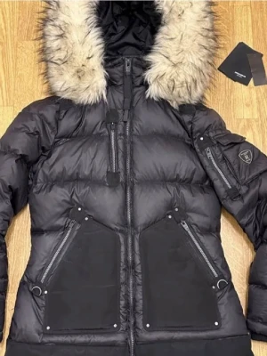 Svart lång dunjacka med pälskrage - Snygg svart dunjacka från Rockandblue med lång passform och stor pälskantad huva. Jackan har flera dragkedjeförsedda fickor, patch på ärmen och quiltad design. Perfekt för kalla vinterdagar och riktigt varm tack vare dunfyllningen. Endast provad