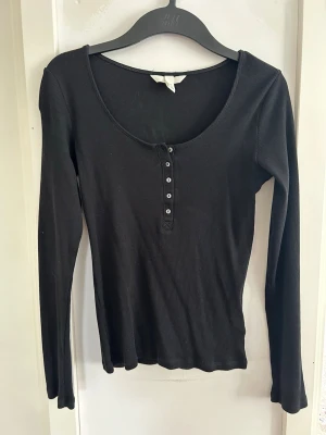 Svart ribbad långärmad topp - En svart ribbad långärmad topp med rund halsringning och knappar framtill. Toppen har en slim passform och är tillverkad i ett mjukt, stretchigt material som sitter snyggt på kroppen. Perfekt att styla med jeans eller kjol.