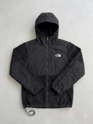 Svart hoodiejacka från The North Face - Svart hoodiejacka från The North Face med dragkedja framtill och justerbar huva. Jackan har en mix av slät och fluffig fleece, två sidofickor och logga på bröstet och ryggen. Perfekt för lager-på-lager och chill stil. Storlek: S