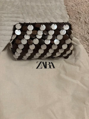 Kuvertväska med paljetter från Zara - Snygg kuvertväska från Zara dekorerad med stora runda paljetter i brunt och pärlemorvitt. Väskan har en guldfärgad kedja som axelrem och stängs upptill. Materialet är en mix av tyg och plast/paljetter för en unik look.