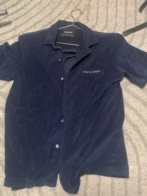Mörkblå sammetsskjorta Lyle & Scott - Snygg mörkblå skjorta från Lyle & Scott i mjuk sammet. Skjortan har korta ärmar, klassisk krage, knappar framtill och en bröstficka med broderad logga. Perfekt för dig som vill ha en stilren men ändå unik look.