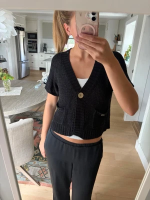 Svart kortärmad kofta  - Svart kortärmad stickad kofta från Flash Woman med korta ärmar, djup V-ringning och två framfickor. Koftan har en knapp framtill och ribbstickade detaljer vid ärmslut och nederkant. Perfekt att slänga över ett linne för en chill look.