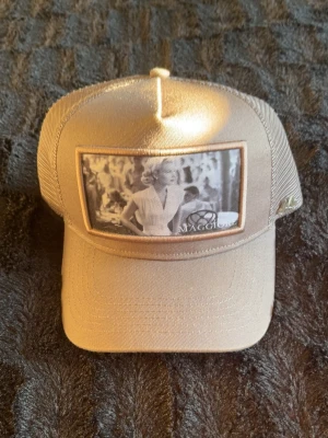 Maggiore beige keps - Snygg beige keps med mesh på sidorna och ett svartvitt retrofoto framtill. Kepsen har böjd skärm och justerbar baksida. Perfekt för dig som gillar unik streetstyle och vintagevibbar. Materialet är en mix av tyg och mesh för extra ventilation.