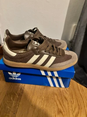 Bruna Adidas sneakers med vita ränder - Säljer ett par bruna Adidas sneakers med klassiska vita ränder på sidan. Skorna har en kombination av mocka och mesh, bruna skosnören och en gummisula i beige. Perfekta för dig som gillar retrostil och streetwear. Använde 2 gånger innan de växte ut 
