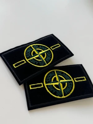 Stone Island tygmärken 2x - Två klassiska tygmärken från Stone Island i svart med broderad gul och grön kompasslogga. Perfekta att sy eller fästa på jackor, väskor eller andra plagg för en streetwear-look. Märkena är rektangulära och har tydliga detaljer.