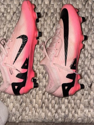 Rosa Nike fotbollsskor - Säljer ett par snygga rosa fotbollsskor från Nike med svart sula och svarta detaljer. Skorna har snörning, låg profil och är tillverkade i ett lätt syntetmaterial. Perfekta för dig som vill sticka ut på planen med stilrena och sportiga skor.