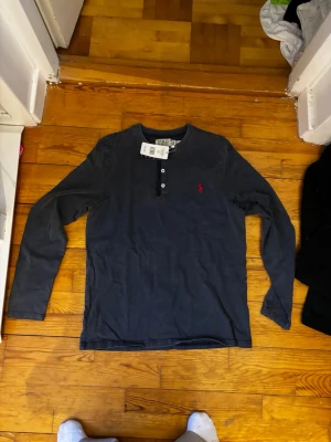 Mörkblå långärmad topp från Polo Ralph Lauren - Snygg mörkblå långärmad topp från Polo Ralph Lauren med klassisk röd broderad logga på bröstet. Toppen har rund halsringning och knappar framtill. Perfekt till jeans eller kjol för en clean och stilren look.