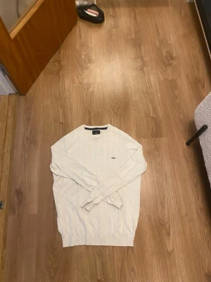 Vit kabelstickad tröja - Säljer en vit kabelstickad tröja med rund hals och diskret broderad logga på bröstet. Tröjan har långa ärmar och ribbade muddar vid ärmslut och nederkant. Perfekt för dig som gillar klassisk och stilren look.