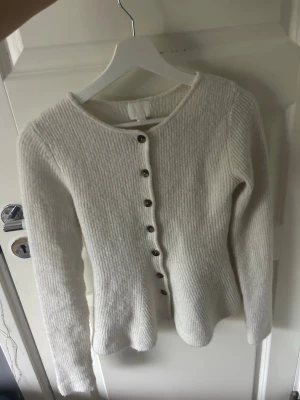 Vit ribbstickad kofta med knappar - Säljer en vit ribbstickad kofta med rund hals och långa ärmar. Koftan har snygga mörka knappar framtill och är gjord i ett mjukt, tjockare material som känns riktigt cozy.