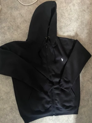 Svart hoodie  - Säljer en svart hoodie med dragkedja från Polo Ralph Lauren. Klassisk design med vit broderad logga på bröstet, huva med dragsko och två fickor framtill. Perfekt för en chill och stilren look. Om du är intresserad är det bara att höra av dig  så kan vi diskutera priset. 