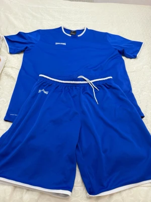 Blått träningsset från Spalding - Säljer ett blått träningsset från Spalding med matchande t-shirt och shorts. Setet har vita detaljer vid hals, ärmslut och benslut samt dragsko i midjan. Tillverkat i lätt och snabbtorkande polyester, perfekt för sport och träning.