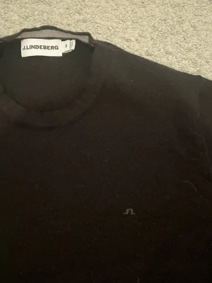 Svart stickad tröja J.Lindeberg - Klassisk svart stickad tröja från J.Lindeberg med rund halsringning och diskret logga på bröstet. Tröjan har lång ärm och är perfekt för lager på lager. Enkel och stilren design som passar till det mesta. Small i storlek men har krympt lite i storlek!! 