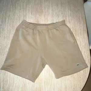 Lacoste Beige shorts  - Snygga beige shorts från Lacoste med elastisk midja och diskret Lacoste-logga på ena benet. Klassisk passform och mjukt material som passar perfekt till sommaren. En bakficka ger extra stilpoäng.