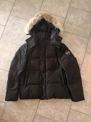 Svart dunjacka med pälskantad huva - Snygg svart dunjacka med avtagbar huva och pälskant. Jackan har en patch på ärmen, dragkedja och knappar framtill samt flera fickor. Perfekt för kalla vinterdagar och riktigt varm tack vare dunfyllningen. Säljer billigt nu innan vintern passa på🫡 gratis retur upp till 14 dagar.