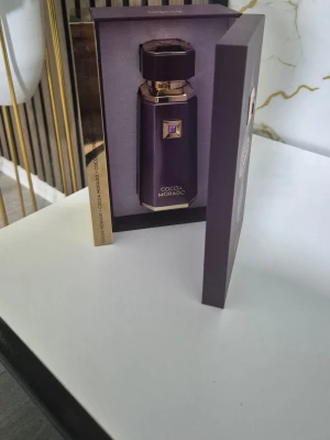 Cocoa Morado lyxig parfymflaska - Elegant parfymflaska i mörklila med gulddetaljer och geometrisk design. Flaskan har en exklusiv känsla och levereras i en matchande ask med guldfärgade kanter. Perfekt för dig som gillar lyxiga och unika dofter.