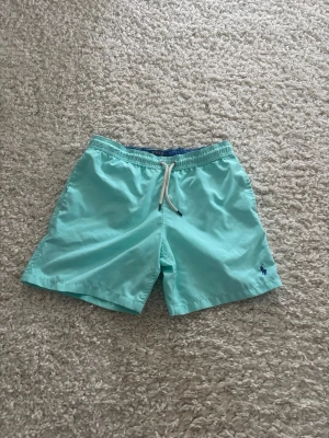 Polo Ralph lauren badbyxor  - Snygga turkosa badbyxor från Polo Ralph Lauren med vit dragsko i midjan och elastisk resår. Klassisk passform, korta ben och liten blå logga broderad framtill. Bakficka med knapp och tillverkade i snabbtorkande material. Klippt bort lapp men har storlek som passar 12 år.