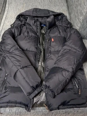 Svart pufferjacka från Polo Ralph Lauren - Priset kan diskuteras                              Snygg svart pufferjacka från Polo Ralph Lauren med röd broderad logga på bröstet. Jackan har huva, dragkedja framtill och två sidofickor med dragkedja. Perfekt för kalla vinterdagar och riktigt skön att bära.