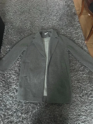Klassisk grå ullkappa - Stilren grå kappa i ullmix med raka linjer och breda slag. Kappan har två fickor framtill och en enkel knäppning. Perfekt för dig som gillar minimalistisk och tidlös stil. Passar till lager-på-lager och funkar lika bra till jeans som till kostymbyxor.
