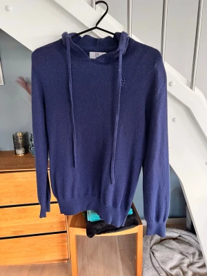 Mörkblå Kashmir hoodie 158/164 - Säljer en mörkblå kashmir och merinoull hoodie med huva från Bvaldi med långa dragsnören. Tröjan har en broderad logga på bröstet och är gjord i ett mjukt, stickat material. Perfekt för dig som gillar en clean och stilren look. Inköpt på KidsBrandStore för 1299kr
