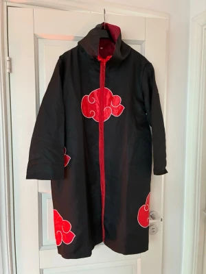 Akatsuki cosplaykappa Naruto - Säljer en svart cosplaykappa inspirerad av Akatsuki från Naruto. Kappan har röda moln med vit kant, hög krage och dragkedja framtill. Perfekt för anime- och cosplayfans som vill sticka ut på event/konvent eller som Halloweenkostym. Tillbehör på bilden ingår
