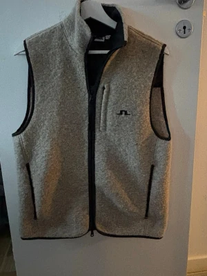 Jlindeberg väst  - Säljer en ljusgrå fleeceväst med svart kantband och hel dragkedja framtill. Västen har två sidofickor med dragkedja och en bröstficka. Materialet är mjukt och fluffigt, perfekt för lager på lager. Liten broderad logga på bröstet.