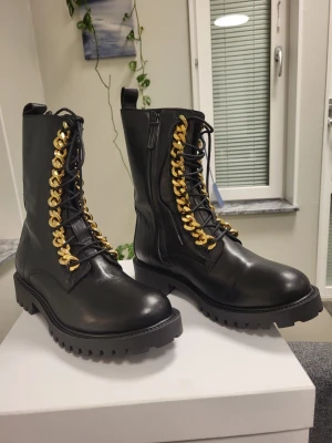 Nya! H&M Moschino Kängor Svarta gulddetalje EUR 43 - Säljer ett par svarta kängor i skinnimitation med chunky sula och snörning dekorerad med grova guldfärgade kedjor. Boots med dragkedja på sidan för enkel påtagning och cool, edgy look. Perfekta för dig som vill sticka ut med detaljer.