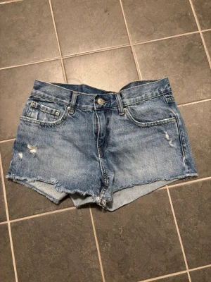 Blå jeansshorts med rå kant - Säljer ett par blå jeansshorts från perfect jeans i storlek 36. Shortsen har råa kanter, slitningar och klassisk femficksmodell. Perfekta för sommaren och ger en avslappnad look med sin slitna denimstil.