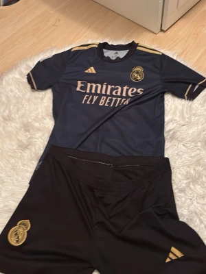 Real Madrid matchställ Benzema #9 - Snyggt Real Madrid matchställ med tröja och shorts i mörkblått och guld. Tröjan har klubbmärke, Adidas-logga och sponsortryck framtill samt Benzema och nummer 9 på ryggen. Tillverkad i lätt och ventilerande material, perfekt för fotbollsträning eller match.