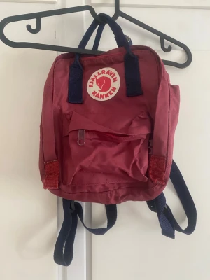 Fjällräven Kånken ryggsäck i - Fjällräven Kånken ryggsäck i vinrött med marinblåa axelremmar och handtag. Ryggsäcken har ett huvudfack med dragkedja, en framficka med dragkedja och två öppna sidofickor. Inuti finns en etikett för namn och kontaktuppgifter.