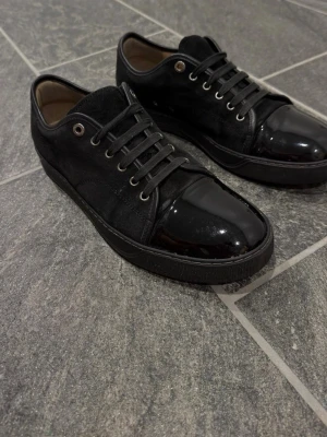 Svarta lanvin - Snygga svarta sneakers med cool kombination av mocka och glansigt lack på tån. Skorna har svarta snören, metallöljetter och låg profil. Perfekta för dig som vill ha en stilren men ändå unik look.