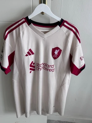 Vit Liverpool matchtröja Adidas - Snygg vit Liverpool matchtröja från Adidas med vinröda detaljer och klubbmärke på bröstet. Tröjan har svarta och vinröda ränder på axlarna, rund halsringning och tryckta sponsorer. Perfekt för dig som älskar fotboll och vill visa ditt stöd för LFC.