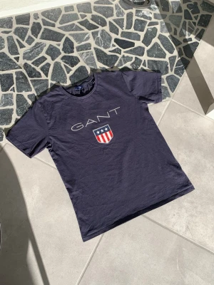 Mörkblå GANT t-shirt med tryck - Snygg mörkblå t-shirt från GANT med stort logotryck och amerikanskinspirerat sköldmärke på bröstet. Klassisk rund hals och korta ärmar. Tillverkad i mjuk bomull som känns skön mot huden.