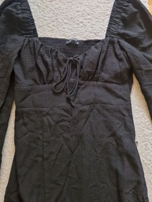 Svart långklänning med puffärm - Snygg svart långklänning med puffiga långärmar och knyt i ringningen. Klänningen har en slits framtill och smock upptill för en bekväm passform. Materialet är strukturerat och ger en trendig look.