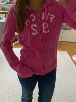 Rosa abercrombie zip up hoodie  - Säljer en rosa hoodie med dragkedja och vita tryckta bokstäver och siffror på bröstet. 