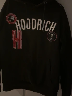 Svart hoodie från Hoodrich - Säljer en svart hoodie från Hoodrich med vita och röda patches och tryck på bröstet. Hoodien har stor huva, dragsnören och känguruficka framtill. Stora bokstäver och coola detaljer gör den perfekt för streetwear-stilen.