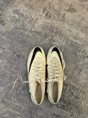 Beige och svarta fotbollsskor - Snygga fotbollsskor i beige med svarta detaljer och snörning. Skorna har en låg profil och är tillverkade i ett lätt syntetmaterial som ger bra känsla på planen. Perfekta för dig som vill ha stilrena och moderna skor till matchen.Fg Nike lemonad inga sulor