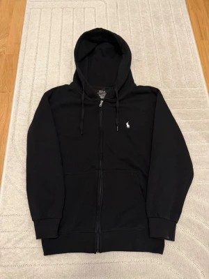Polo Ralph Lauren zip hoodie - Säljer en svart hoodie från Polo Ralph Lauren i custom slim fit. Hoodien har dragkedja, huva med snören  och en liten vit logga på bröstet. Perfekt för en chill och stilren look.