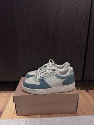 Vita och ljusblå sneakers - Snygga sneakers i vitt och ljusblått med klassisk låg profil och vita skosnören. Ovandelen är i en mix av mocka och textil, vilket ger en stilren och fräsch look. Perfekta för dig som gillar enkel men trendig stil.